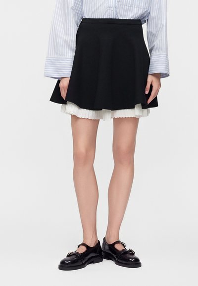 Femme portant une jupe noire évasée avec une sous-couche plissée blanche, une chemise rayée à manches longues et des chaussures en cuir noires à boucle, debout sur fond blanc.