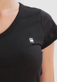 Czarny bawełniany t-shirt z dekoltem w serek i krótkimi rękawami. Posiada małe białe logo na lewej piersi. Gładka tekstura, podstawowy design.