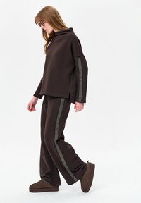 Bruin loungewear-set met een capuchontrui met zijinkepingen en een patroonbijlage, gecombineerd met bijpassende losse broek en pantoffels.