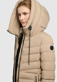 Beige pufferjack met een grote capuchon, gewatteerde textuur, rits aan de voorkant en trekkoordaccenten. Heeft een ronde logopatch op de mouw.