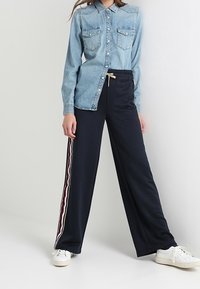 Džínová košile se dvěma náprsními kapsami a zapínáním na knoflíky, doplněná navy širokými kalhotami s červenými a bílými bočními pruhy a stahovací šňůrkou.