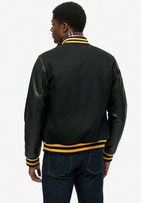 Zwarte varsityjack met leren mouwen, gele en zwarte gestreepte ribgebreide manchetten en tailleband, afgewerkt met een geruite kraagdetail.