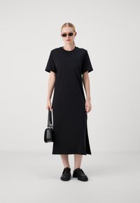 Black t-shirt dress with short sleeves and side slits, made from a smooth fabric. Model carries a small black handbag and wears black shoes.