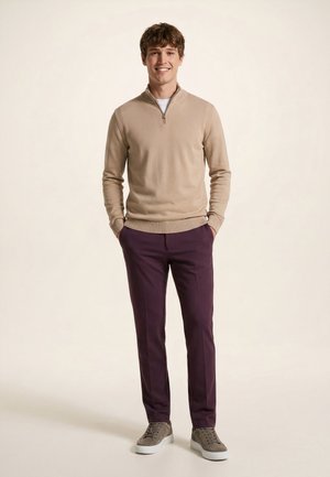 Egy fiatal férfi áll, mosolyog, bézs cipzáras pulóvert, lila nadrágot és szürke tornacipőt visel, kezei a zsebében, egyszínű világos háttér.