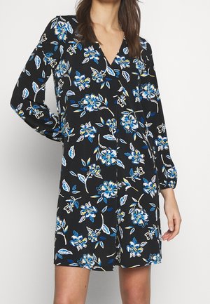 Femme portant une robe noire à manches longues avec un motif floral bleu et blanc, debout devant un fond blanc uni.