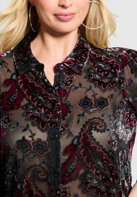 Blusa nera trasparente con motivi paisley e floreali in rosso, blu e mauve, caratterizzata da un colletto e chiusura con bottoni. Orecchini a cerchio argentati visibili.