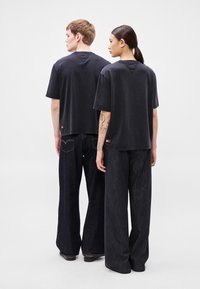Tricouri negre supradimensionate cu pantaloni largi din denim închis la culoare, cu design minimalist și detalii contrastante de cusături, văzute din spate.