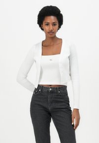 Hvitt langermet cardigan med knappeåpning, over en hvit kort tanktop. Kombinert med høy-hvite svarte jeans, med tettsittende passform.