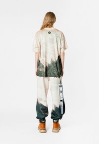 Camisa de gran tamaño y manga corta en beige con un estampado de montañas en verde oscuro. Combinada con pantalones color crema que presentan un diseño similar y etiquetas. Botas marrones.