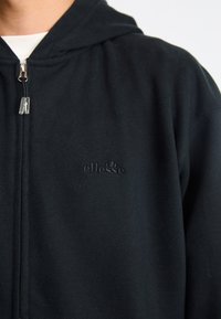 Čierna mikina na zips vyrobená z mäkkej látky s významným vyšívaným logom na hrudi. Obsahuje kapucňu na šnúrku a má štandardný strih.
