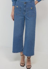 Jeans in denim blu a gamba larga con due grandi tasche frontali, bottoni in ottone, vita alta e orli rifiniti con cura.