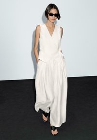 Haut sans manches en lin blanc avec décolleté en V et ourlet asymétrique, associé à un pantalon blanc à jambe large. Modèle portant des lunettes de soleil noires et des sandales.