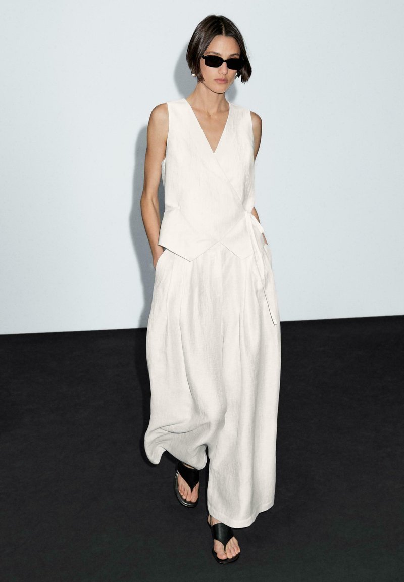 Haut sans manches en lin blanc avec décolleté en V et ourlet asymétrique, associé à un pantalon blanc à jambe large. Modèle portant des lunettes de soleil noires et des sandales.