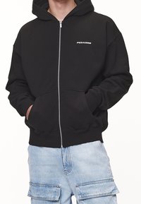 Musta vetoketjullinen huppari, valmistettu puuvillasta, jossa on huppu, etutaskut, resorit hihansuissa ja pieni valkoinen "PEGADOR"-logo rinnassa.