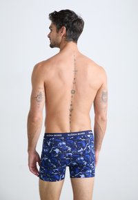 Man with tattoos on back and arms wearing blue patterned Björn Borg boxer briefs, standing against a plain light background.