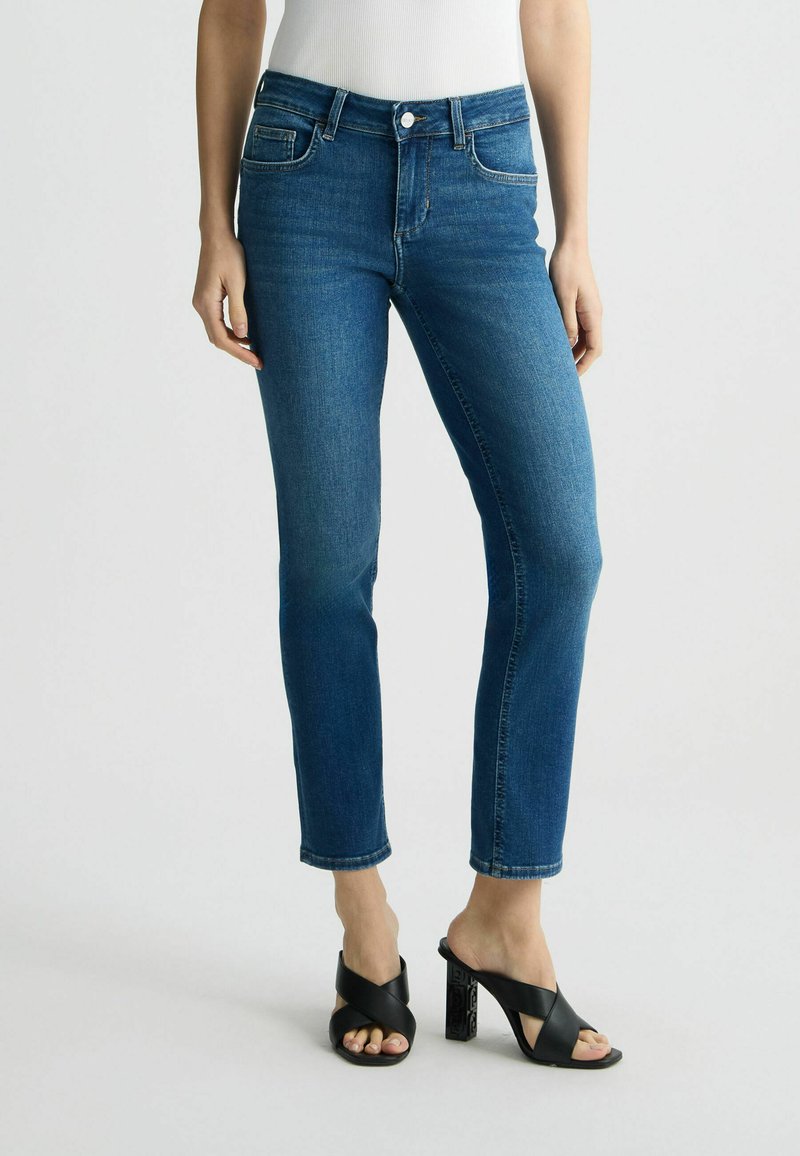 Jeans de denim azul oscuro con un corte slim, cinco bolsillos y cierre frontal de botón. Presenta una pierna ligeramente recortada con un dobladillo limpio.