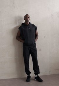 Man in een mouwloos zwart Puma hoodie, zwarte sweatpants en zwarte sneakers, staande tegen een lichtgrijze tegelwand met handen in de zakken.