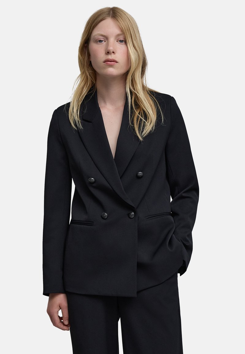 Jong persoon met lang blond haar draagt een zwarte dubbelzijdige blazer en bijpassende broek, staand met één hand in de zak.