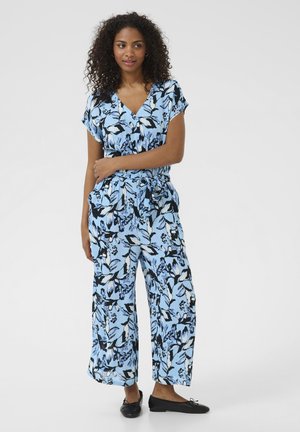Mono floral azul claro con mangas cortas, piernas anchas, patrones de hojas en blanco y negro, y un detalle de cintura anudada. Unos zapatos planos negros completan el look.