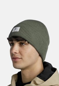 Gorro de punto verde oliva con textura acanalada y una etiqueta de tela blanca con un logotipo. Material suave, diseño ajustado y borde doblado.