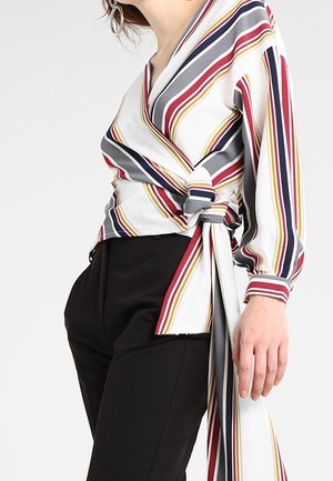 Woman wearing a long-sleeve, wrap-style blouse with red, yellow, gray, and navy stripes, tied at the waist, paired with black pants.