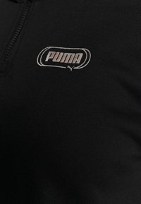 Camiseta atlética negra con cuello de cremallera de cuarto. Presenta un logo plateado de Puma en el área del pecho izquierdo. Tejido suave y texturizado.