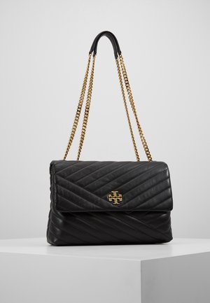 Zwarte gewatteerde leren handtas met gouden kettingriemen en een gouden logo-sluiting, tentoongesteld op een wit voetstuk tegen een grijze achtergrond.