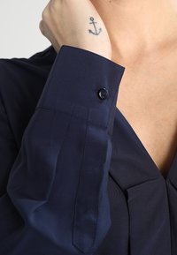Mornarica plava košulja s dugmetom na manžetama, izrađena od glatkog materijala i s V-izrezom. Manžeta sadrži detalj crnog dugmeta.