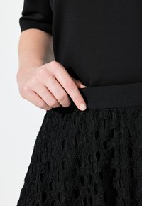 Mustapuku, jossa on sileä vartalo-osa ja teksturoitu hame, jossa on pyöreitä aukko-kuvioita. Käsi koskettaa vyötäröä säätämistä varten.