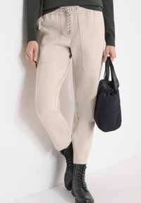 Pantalon beige clair fuselé avec poches avant et taille à cordon, accompagné d'un sac à main noir matelassé et de bottes noires à lacets.