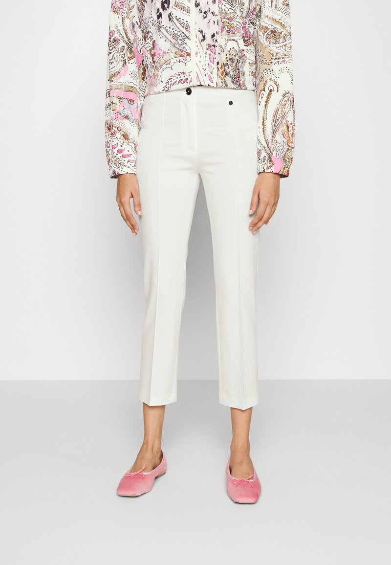 Pantaloni bianchi a vita alta con taglio sartoriale e cuciture frontali, abbinati a scarpe slip-on in velluto rosa, su uno sfondo neutro.