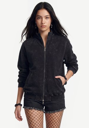 Vrouw met lang donker haar die een zwart jack met ritssluiting draagt, zwarte denimshorts met rafelige randen en visnetkousen, staand tegen een witte achtergrond.