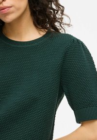 Jersey de color verde oscuro de punto, con un patrón texturizado, mangas cortas y cuello de canalé. Tela suave con un ajuste ceñido.