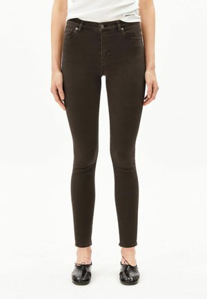 Persoon draagt een donkerblauwe skinny jeans met hoge taille, een witte mouwloze top en zwarte ballerina's, staand tegen een effen witte achtergrond.