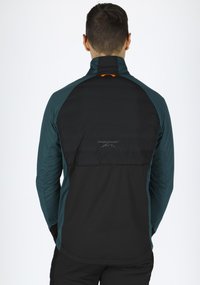 Let vægtkappe i teal og sort, med høj krave, lynlåslomme på bagsiden og logodetalje på den øverste del af ryggen.