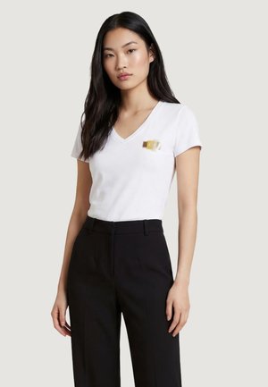 Donna con capelli lunghi e scuri, che indossa una maglietta bianca a V e pantaloni neri a vita alta su uno sfondo chiaro e semplice.