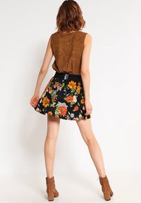 Brauner Wildleder-Ärmellos-Top kombiniert mit einem schwarzen Rock mit bunten Blumenmustern. Klobige braune Stiefeletten runden den Look ab.