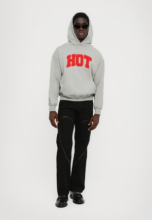 Sweat à capuche gris avec le mot "HOT" en lettres rouges, pantalon noir avec des accents d'étoiles dorées, et chaussures noires brillantes. Le modèle porte des lunettes de soleil.