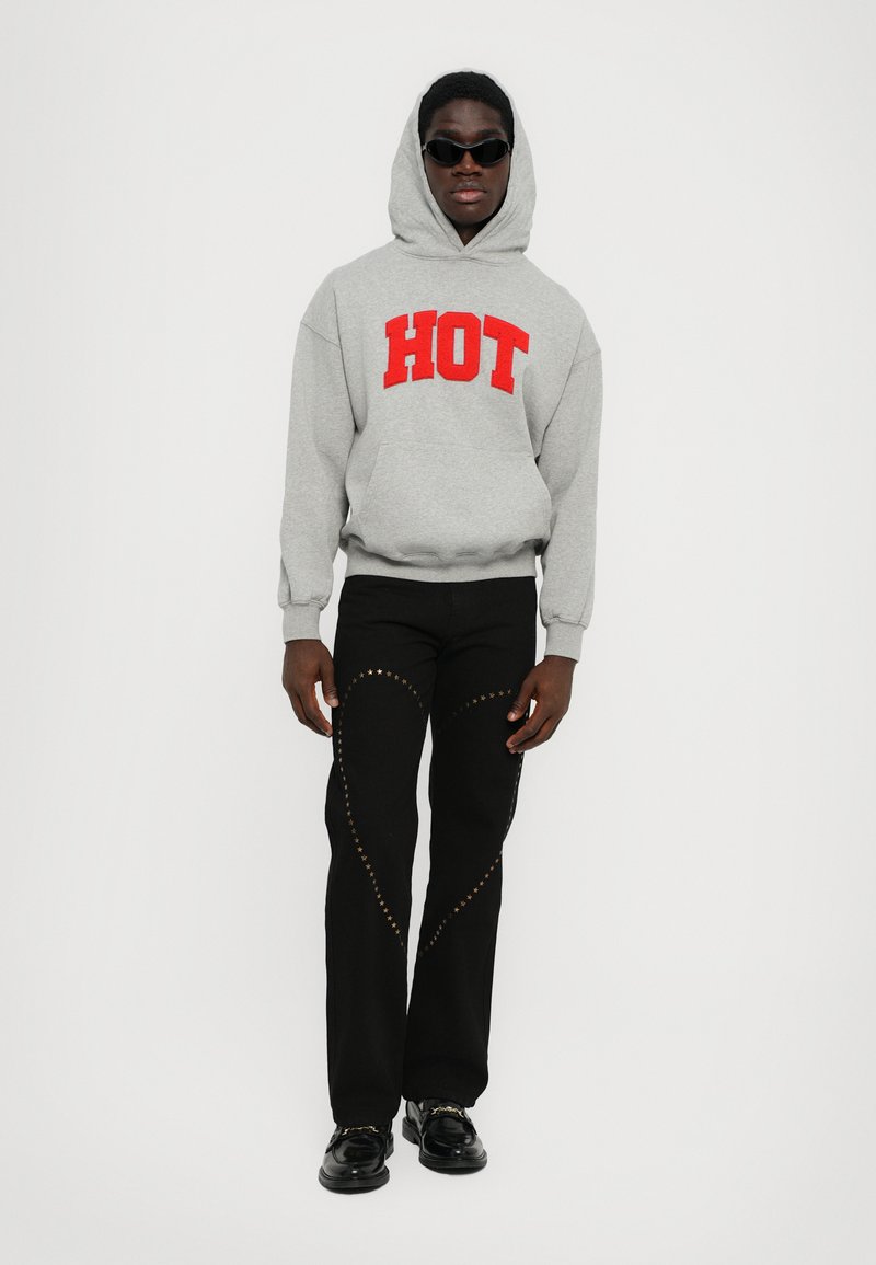Sweat à capuche gris avec le mot "HOT" en lettres rouges, pantalon noir avec des accents d'étoiles dorées, et chaussures noires brillantes. Le modèle porte des lunettes de soleil.