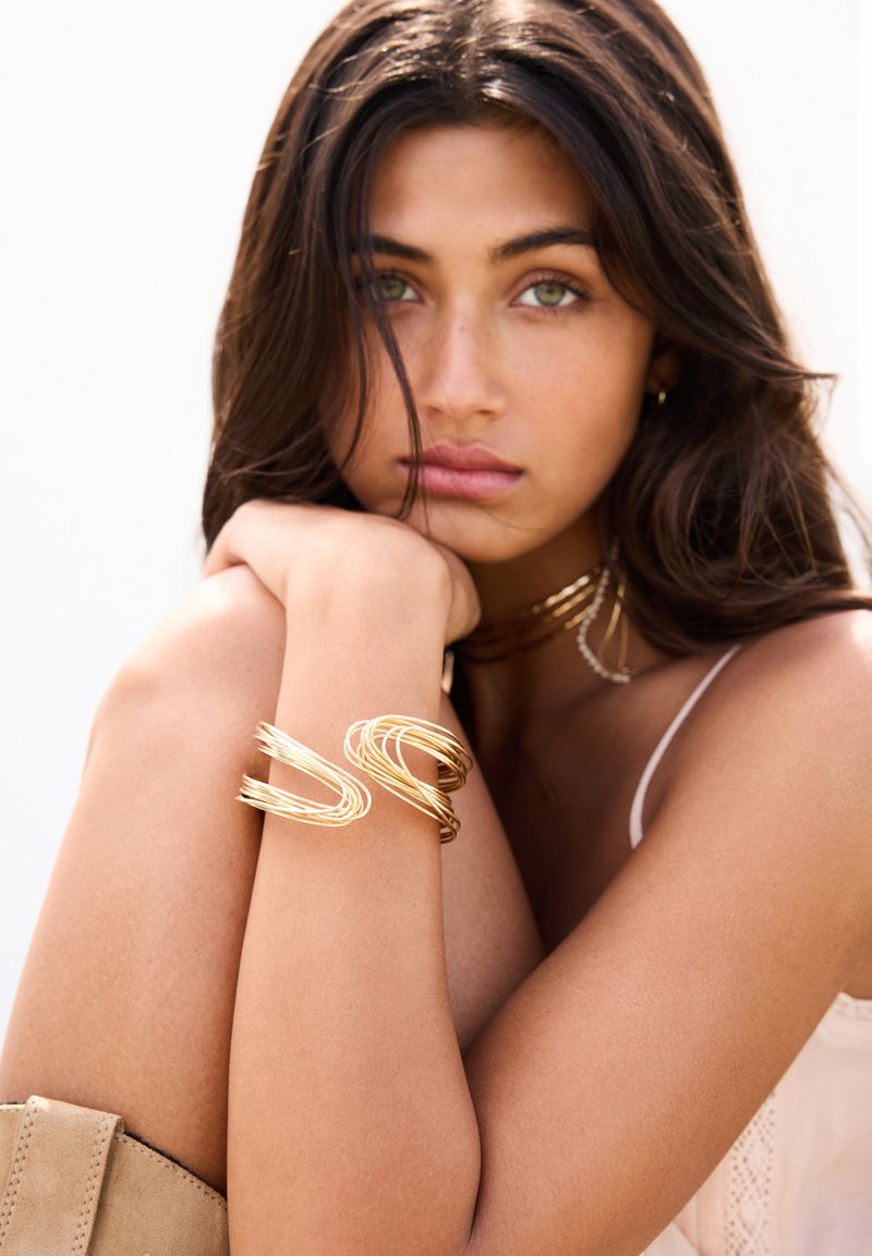 Bracciale in metallo dorato con un design ondulato. Texture liscia e finitura lucida. Modello che indossa una maglietta chiara, con i capelli che scendono.