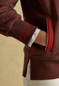 Hnedá mikina so ceruzkovými manžetami a červenými pruhmi. Pozdĺžny rozparok pre väčší komfort. Na manžete je viditeľná biela košeľa, ktorá kontrastuje s hnedou.