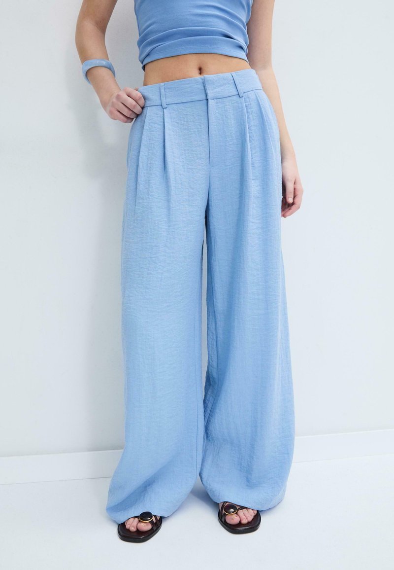 Femme portant un pantalon large bleu clair, un crop top assorti, des sandales noires et un bracelet bleu, debout contre un mur blanc.