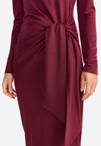 Bordeauxfarbenes Kleid mit langen Ärmeln und einem gedrehten Knoten-Detail an der Taille, aus einem weichen, strukturierten Stoff und mit einer eleganten Silhouette.