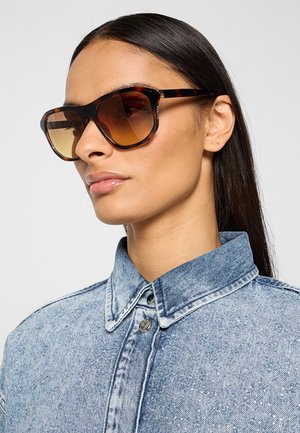 Donna con lunghi capelli scuri indossa grandi occhiali da sole tartarugati e una camicia di jeans azzurro chiaro con bottoni su uno sfondo semplice.