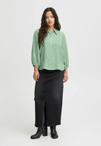 Hele rohelise kaelusega pluus, millel on puhv-sleeved, kombineeritud musta denimpika seelikuga, millel on eeslõige ja kantud mustade pahkluudega saapadega.