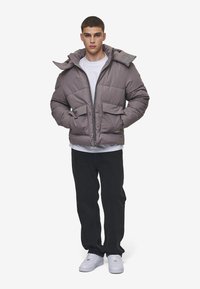 Chaqueta tipo puff en gris claro, con cierre frontal, dos bolsillos delanteros y capucha. Lleva puesta sobre una camisa blanca y combinada con pantalones negros.
