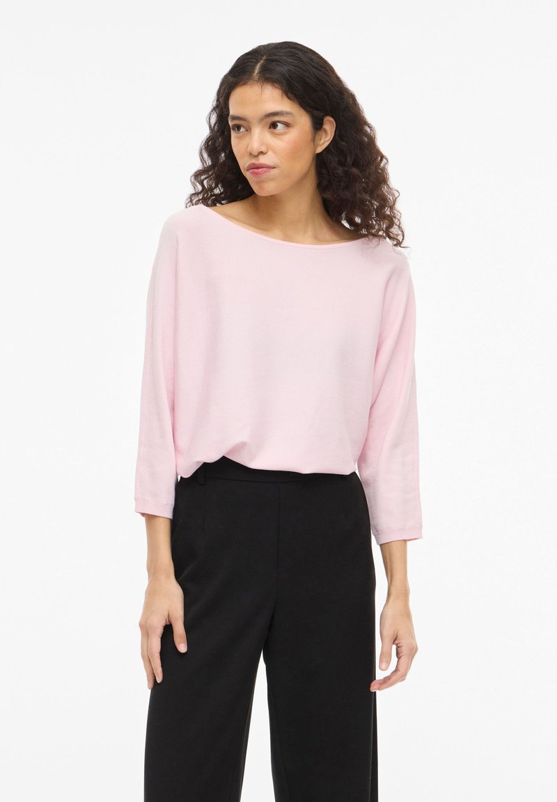 Sieviete ar cirtainiem matiem, tērpusies vaļīgā gaiši rozā garroku krekliņā un melnās augstu jostasvietu plaša gurna biksēs, stāvot pret vienkāršu fonu.