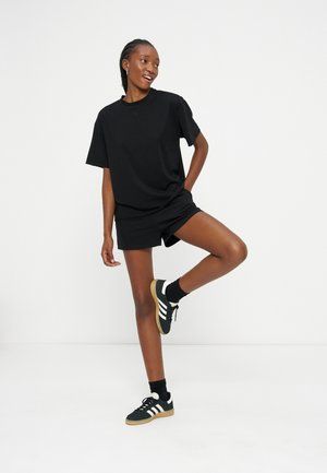 Woman in black oversized t-shirt and shorts, black Adidas sneakers, lifting one leg, smiling against plain white background.
