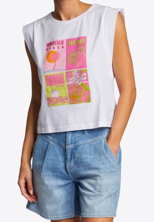 Top court sans manches blanc avec un motif rétro hawaïen coloré, associé à un short en jean bleu clair, une main dans la poche.