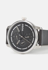 Schwarze analoge Uhr mit einem runden silbernen Gehäuse, drei Subdials, minimalistischen Design und einem Lederarmband. Markenname "HUGO" auf dem Zifferblatt angezeigt.